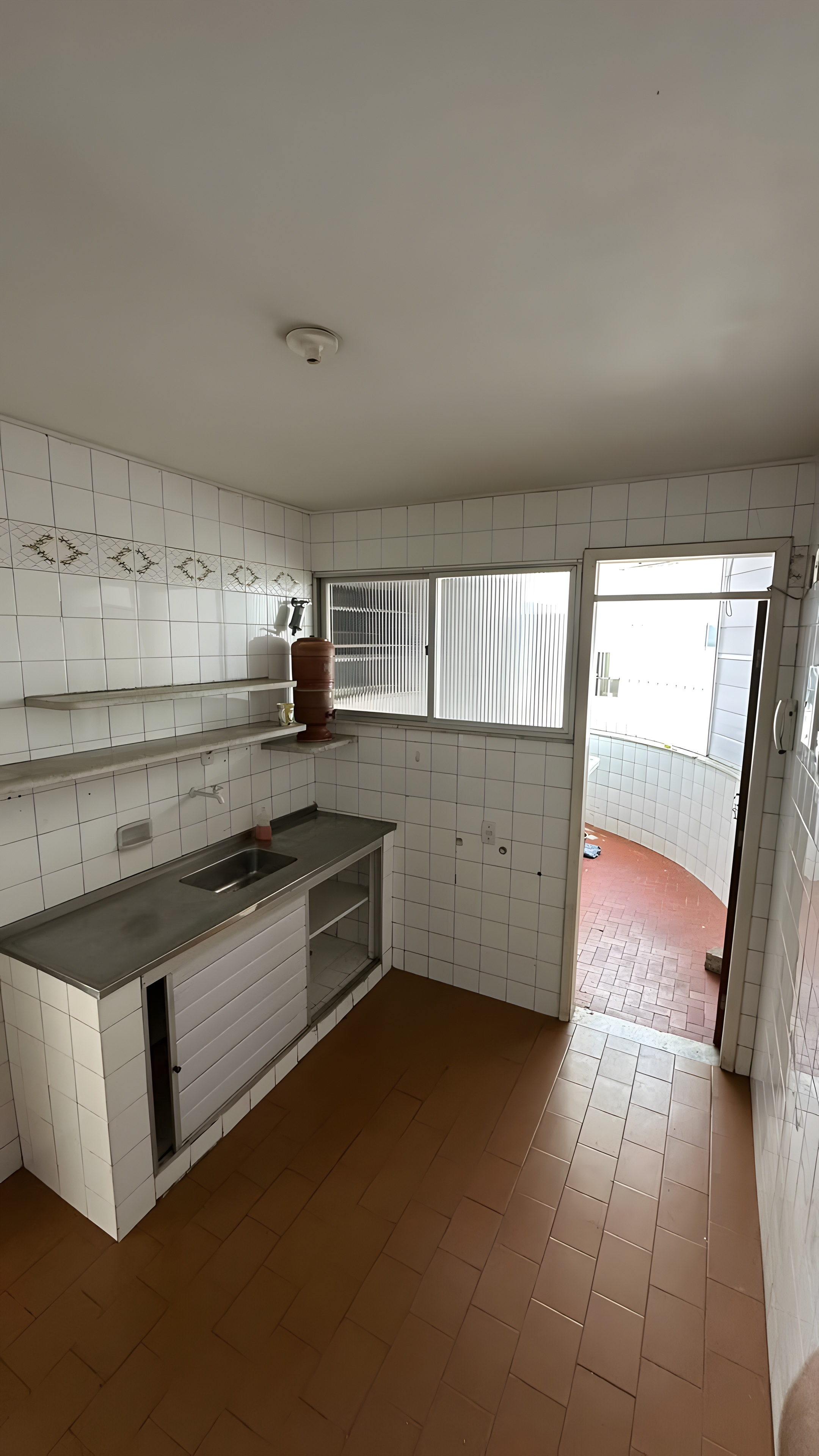 Cozinha original com azulejos brancos, bancada em inox e área de serviço ao fundo